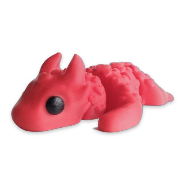 articulated-love-dragon-fidget-toy-poseable-desk-buddy-1 A pink articulated dragon figure covered in heart-shaped scales, lying on a white surface with black eyes visible.