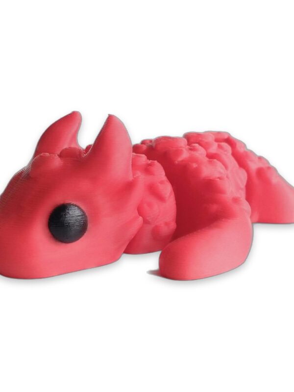 articulated-love-dragon-fidget-toy-poseable-desk-buddy-1 A pink articulated dragon figure covered in heart-shaped scales, lying on a white surface with black eyes visible.